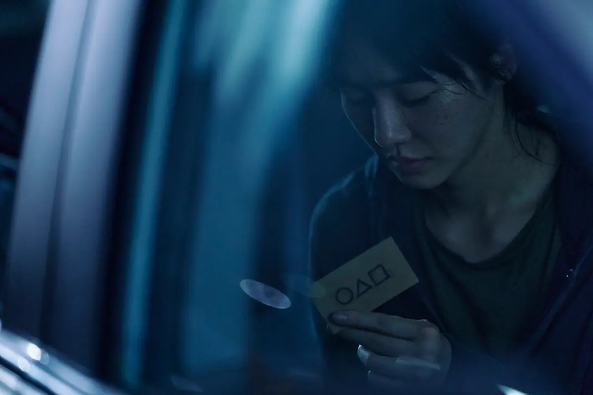 Reflection in a car window of a woman looking at a small card with a circle, triangle, and square on it in Season 2 of ‘Squid Game’